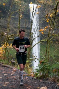 two people running in front of a waterfall on a trail with trees and leaves around them