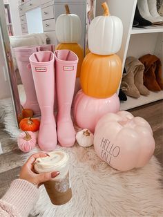 a woman holding a cup in her hand next to boots and pumpkins on the floor