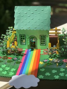 a paper model of a house with a rainbow in the front yard and trees behind it