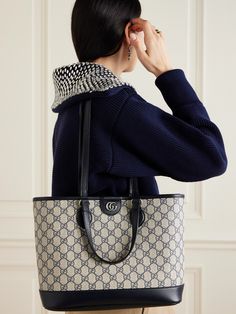 a woman wearing a blue sweater and black pants holding a white gucci tote bag