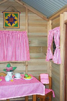 a small cabin with a table and chairs in front of the window, covered by pink gingham curtains