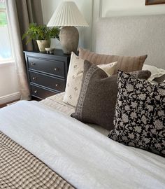 a bed with several pillows on it and a lamp next to the headboard, in front of a window