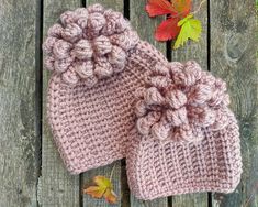 two pink knitted hats with flowers on them sitting on a wooden surface next to autumn leaves