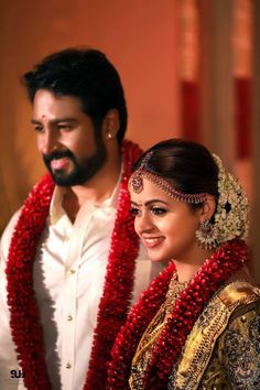 a man and woman standing next to each other in front of a red flower garland