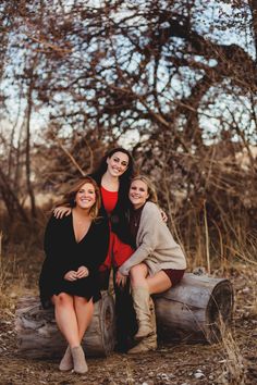 three women are sitting on a log in the woods and posing for a photo with their arms around each other