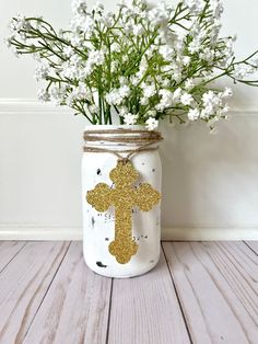 a mason jar filled with white flowers and a gold cross painted on the front is sitting on a wooden table