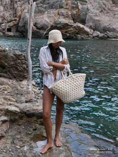 a woman standing on rocks next to water holding a white bag and wearing a straw hat