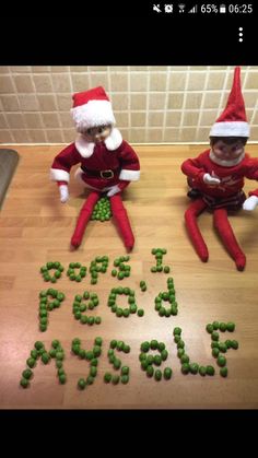 two elfs sitting on top of a wooden table next to green peas and the words i love santa spelled by them