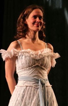 a woman in a white dress standing on stage with her hands behind her back and smiling at the camera