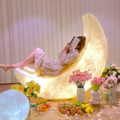 a woman in white dress laying on the moon shaped bed with flowers and petals around it