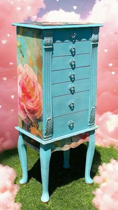 a blue painted dresser sitting on top of a lush green grass covered field with pink clouds in the background