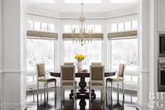 a dining room table with chairs and a vase on top of it in front of two windows