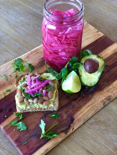 a wooden cutting board topped with food next to a jar of pickles and an avocado