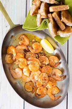 a pan filled with cooked shrimp next to bread and lemon wedges on a wooden table