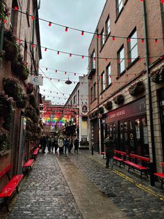 people are walking down an alley way with red benches and lights strung across the street