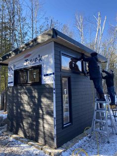 two men are painting the side of a small building in the snow, while another man stands on a ladder