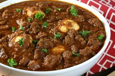 a white bowl filled with meat and potatoes on top of a red table cloth next to a wooden spoon