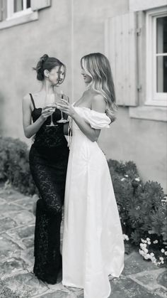 two women standing next to each other in front of a building and holding wine glasses