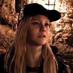 a woman with long blonde hair wearing a black hat and leather jacket standing in front of a stone wall