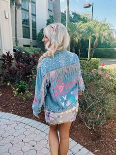 a woman wearing a denim jacket with the word love painted on it, walking down a sidewalk