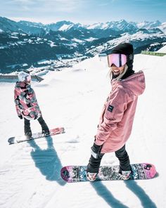 two snowboarders are standing in the snow on top of a hill with mountains behind them