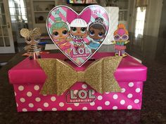 a pink box with gold glitter bow and lol surprisers on the top, sitting on a table