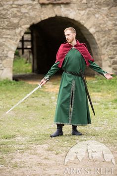 a man dressed in green and red holds two swords while standing on the grass near an old stone building