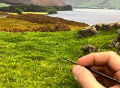 a hand holding a pair of scissors in front of a painting of grass and mountains