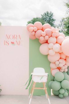 a chair sitting in front of a wall with balloons attached to it and a sign that says nora is one