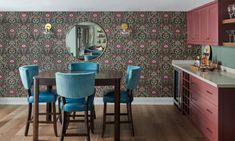 a dining room table with blue chairs in front of a wallpapered kitchen area