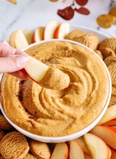 a hand dipping an apple slice into a bowl of peanut butter and apples on the side