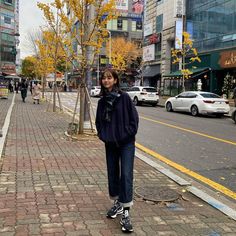 a woman standing on the side of a street next to a tree with yellow leaves