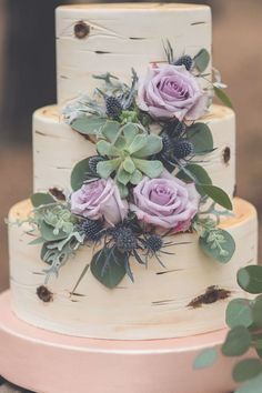 a three tiered cake with purple flowers and greenery on the top is sitting on a wooden table