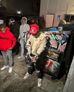 several young men standing around in front of a graffiti covered building with one man wearing a red hoodie