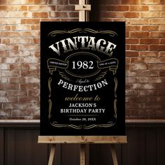 a black and gold birthday party sign on an easel in front of a brick wall