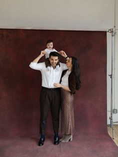 a man and woman holding a baby in front of a red background with a wall behind them