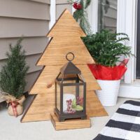 a small wooden christmas tree next to a potted plant on the front porch with a lantern