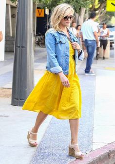 a woman in a yellow dress and jean jacket walking down the street with her hand on her hip