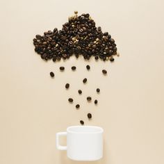 coffee beans falling into a white cup on a beige surface next to a pile of brown coffee beans