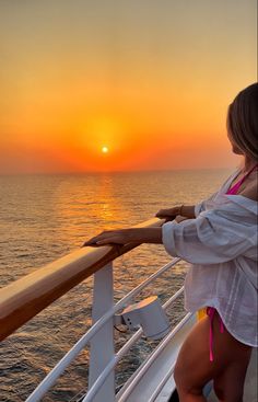 a woman standing on the deck of a boat watching the sun go down over the ocean