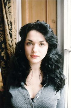 a woman with long black hair standing in front of a window looking at the camera