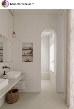 a white bathroom with two sinks and an arched doorway leading to another room in the background