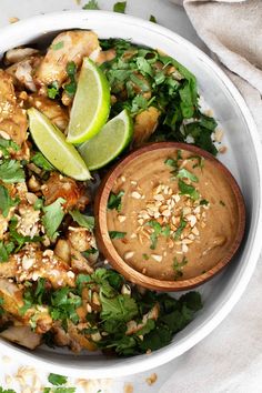 a white bowl filled with chicken and garnished with cilantro, lime wedges and peanuts