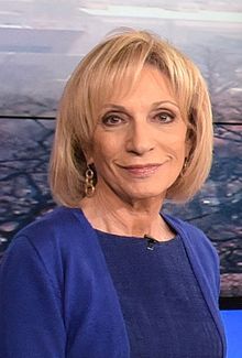 a woman with blonde hair wearing a blue top and gold earrings smiling at the camera