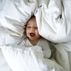 a baby is laying in bed with white sheets on it's head and smiling