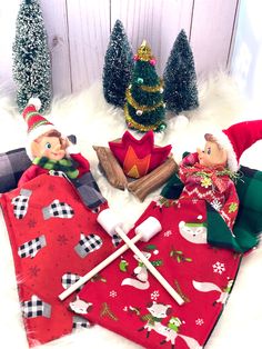 two elfs sitting on top of a red blanket next to christmas trees and other decorations