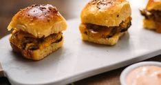 three mini sandwiches sitting on top of a white platter next to dipping sauces