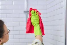 a woman in green and pink gloves is cleaning a shower wall with a rag on it