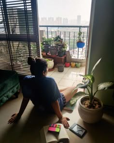 a woman sitting on the floor reading a book next to a window with potted plants