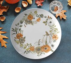 a white plate with orange and yellow flowers on it next to autumn leaves, pumpkins and acorns